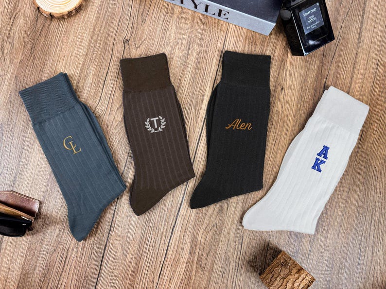 May include: Four pairs of ribbed dress socks in various colors: teal, brown, black, and white. Each sock features embroidered initials or a name in a contrasting color. The socks are arranged on a wooden surface.