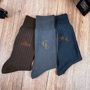 May include: Three pairs of ribbed dress socks in brown, blue-grey, and navy blue. Each sock features embroidered initials or a name in a gold color. The socks are displayed on a wooden surface.