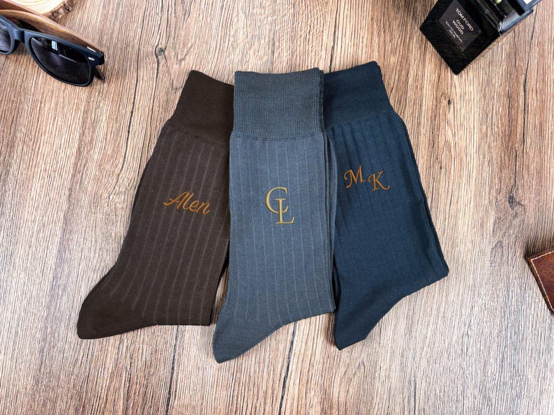 May include: Three pairs of ribbed dress socks in brown, blue-grey, and navy blue. Each sock features embroidered initials or a name in a gold color. The socks are displayed on a wooden surface.