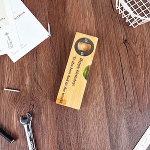 May include: A wooden bottle opener with a metal opener and a level. The opener has the text "Happy birthday! To the best dad in the world" engraved on it. The opener is on a wooden surface.