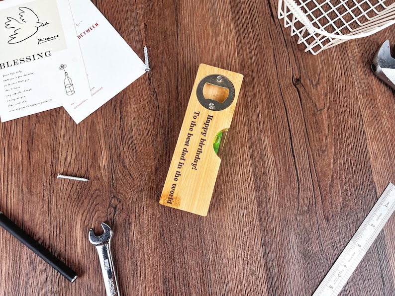 May include: A wooden bottle opener with a metal opener and a level. The opener has the text "Happy birthday! To the best dad in the world" engraved on it. The opener is on a wooden surface.