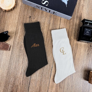 May include: A pair of personalized socks, one black and one white, with embroidered initials. The black sock has the name "Alen" in gold thread, while the white sock features the initials "CL". Sunglasses and a bottle of cologne are also visible.