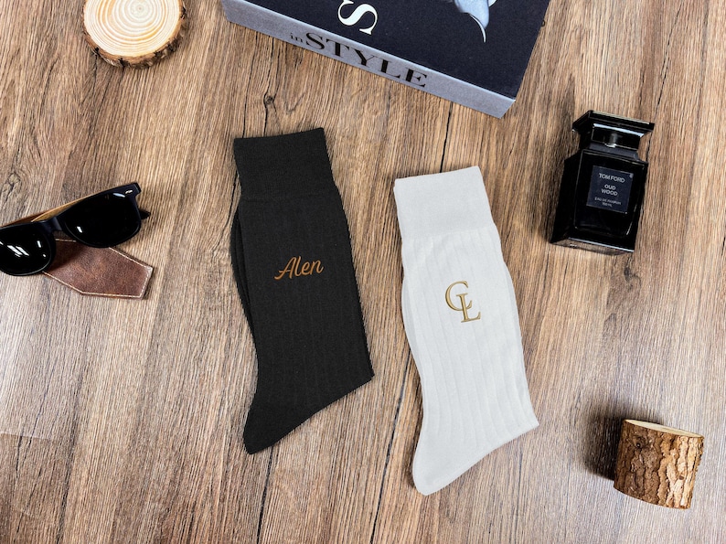 May include: A pair of personalized socks, one black and one white, with embroidered initials. The black sock has the name "Alen" in gold thread, while the white sock features the initials "CL". Sunglasses and a bottle of cologne are also visible.