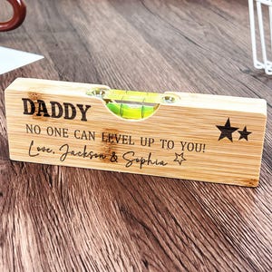 Puede incluir: Un nivel de madera con la palabra "DADDY" grabada, junto con el mensaje "NO ONE CAN LEVEL UP TO YOU! Love, Jackson & Sophia." El nivel tiene una burbuja y gráficos de estrellas.