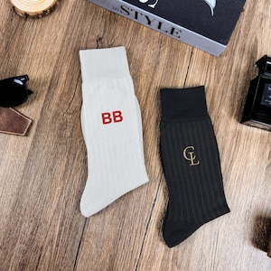May include: A pair of socks, one white with red "BB" embroidery and the other black with a gold "CL" embroidery. The socks are ribbed and sit on a wooden surface, next to sunglasses and a bottle of cologne.