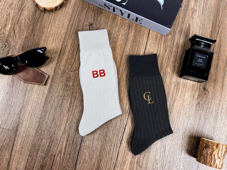 May include: A pair of socks, one white with red "BB" embroidery and the other black with a gold "CL" embroidery. The socks are ribbed and sit on a wooden surface, next to sunglasses and a bottle of cologne.