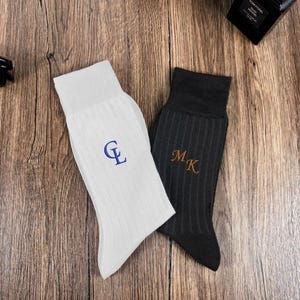 May include: Two pairs of ribbed socks, one white and one dark gray, each embroidered with initials. The white sock features blue initials, while the gray sock has gold initials. The socks are displayed on a wooden surface.