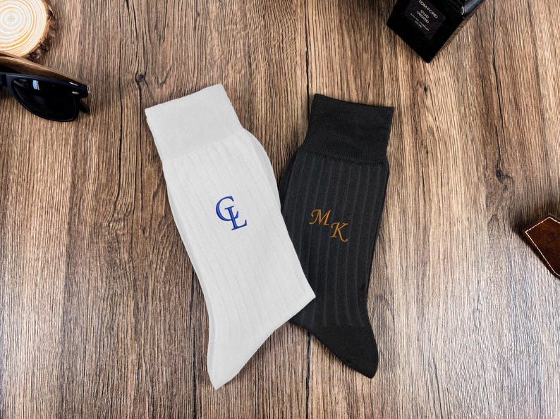 May include: Two pairs of ribbed socks, one white and one dark gray, each embroidered with initials. The white sock features blue initials, while the gray sock has gold initials. The socks are displayed on a wooden surface.