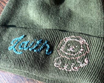May include: Green knit beanie with the word "Faith" embroidered in blue script. A small lion face is embroidered in pink and brown thread. The beanie is made of a ribbed knit fabric.