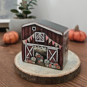May include: A hand-painted, miniature barn with a dark red exterior, a black roof, and a decorative banner. The barn features open doors revealing pumpkins and flowers. It sits on a wooden slice, with pumpkins in the background.