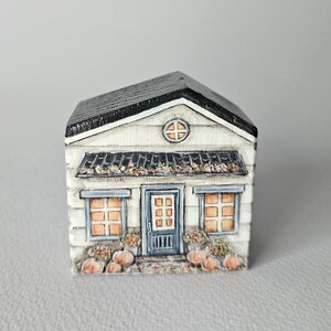 Könnte beinhalten: Ein handbemaltes Miniaturhaus mit schwarzem Dach, weißer Verkleidung und einer blauen Tür. Das Haus hat zwei Fenster mit orangefarbenen Rahmen, ein kleines rundes Fenster und Kürbisse. Das Haus ist ein Dekorationsartikel.