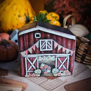 Könnte beinhalten: Eine handbemalte, kleine rote Scheune mit weißem Rand, einem schwarzen Dach und einem Wimpelbanner. Die Scheunentore sind geöffnet und zeigen Kürbisse und Blumen. Das Gesamtthema ist Herbst und Ernte.