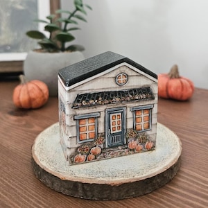 Könnte beinhalten: Ein handbemaltes Miniaturhaus mit schwarzem Dach, weißer Verkleidung und einer blauen Tür. Das Haus ist mit Kürbissen und Herbstlaub dekoriert. Es steht auf einer Holzscheibe, mit Kürbissen im Hintergrund. Eine charmante Herbstdekoration.