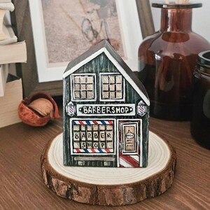 Peut inclure: Un petit bloc de bois peint à la main en forme de bâtiment, représentant un salon de coiffure à l'extérieur vert foncé. La façade présente deux fenêtres supérieures, une vitrine avec des rayures rouges, blanches et bleues et une porte.