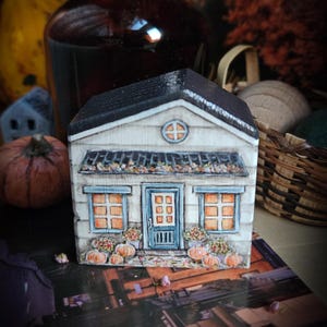 Könnte beinhalten: Ein Miniatur-Holzhaus mit schwarzem Dach, weißer Verkleidung und blauen Zierleisten. Das Haus verfügt über eine Eingangstür, Fenster und eine kleine Veranda, die mit Kürbissen und Blumen geschmückt ist. Eine charmante Herbstdekoration.