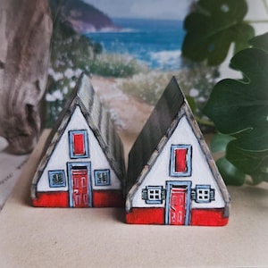 May include: Two miniature, hand-painted wooden houses with white walls, red doors and window frames, and gray roofs. The houses have small windows and shutters. The background shows a coastal scene with blue water and green foliage.
