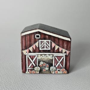 Könnte beinhalten: Eine handbemalte Miniatur-Scheune mit dunkelroter Außenfarbe, schwarzem Dach und weißem Zierrat. Die Scheunentore sind geöffnet und zeigen Kürbisse und Blumengestecke. Ein Banner aus rosa und weißen Dreiecken hängt über den Türen.