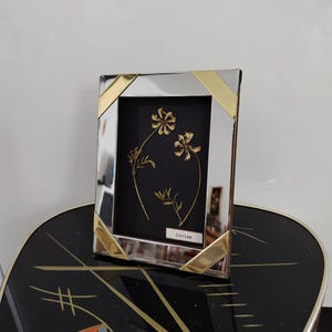 May include: A rectangular silver and gold picture frame with a black background. The frame holds a botanical arrangement of dried flowers. The flowers are a light tan color. The word "Rovien" is printed on a small white label.