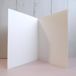 May include: A blank, off-white greeting card standing upright in an open position. The card features a smooth, matte texture and is constructed from heavy cardstock. The interior and exterior surfaces are completely plain, showing no text or graphics.