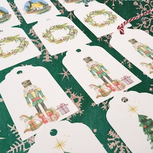 May include: White gift tags with various watercolor illustrations, including a nutcracker, wreaths, and a Christmas tree. The tags are arranged on a green surface with snowflake and star patterns.