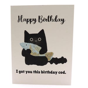 May include: A birthday card with the text "Happy Birthday" and "I got you this birthday cod." A black cat illustration holds a fish. The card is on a cream background.