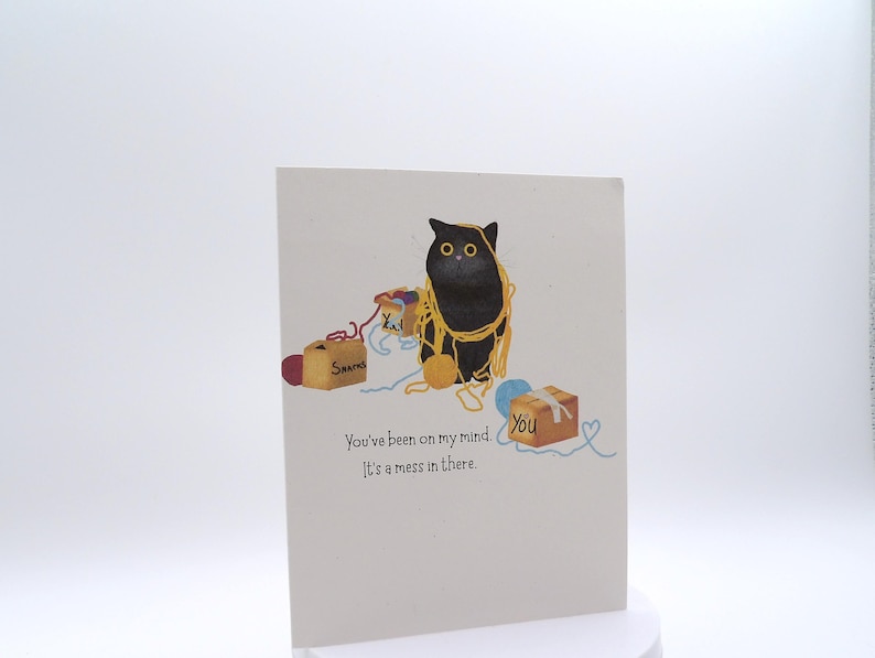 May include: A greeting card featuring a black cat tangled in yellow yarn, surrounded by small cardboard boxes labeled "Snacks" and "You". The text reads, "You've been on my mind. It's a mess in there." The background is a plain, light off-white color.