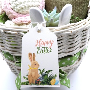 May include: A white wicker Easter basket filled with a pink and white crocheted item, a green plant, and white bunny ears. A tag with a watercolor bunny and the words "Happy Easter" hangs from the basket.