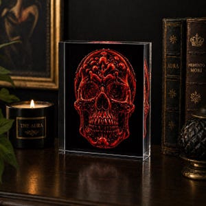 May include: A red skull design encased in a clear, rectangular block. The skull has intricate details and sits against a black background. The block is displayed on a dark wooden surface, with a lit candle and antique books in the background.