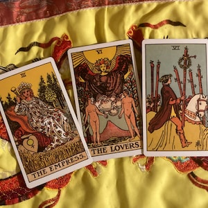 Puede incluir: Se muestran tres cartas del tarot, con ilustraciones detalladas y texto. Las cartas incluyen "The Empress", "The Lovers" y una carta con una persona a caballo. Las cartas están sobre un fondo amarillo y rojo estampado.