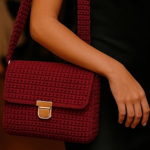 May include: A burgundy crochet shoulder bag with a rectangular shape and a flap closure. The bag features a gold-colored clasp and a matching burgundy strap. The bag is worn over the shoulder.