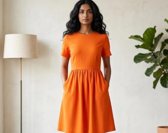 May include: A bright orange dress with short sleeves and a gathered waist. The dress has pockets and falls to the knees. The model is standing in front of a neutral wall with a floor lamp and a potted plant.