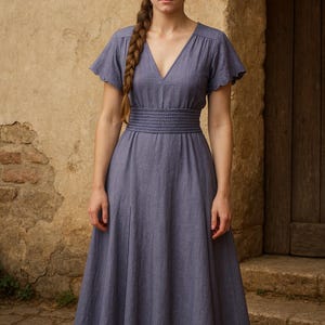 Könnte beinhalten: Ein langes, periwinklefarbenes Kleid mit V-Ausschnitt und kurzen, gewellten Ärmeln. Das Kleid hat eine geraffte Taille und einen fließenden Rock. Das Model hat einen langen Zopf. Der Hintergrund umfasst eine Steinmauer und eine Holztür.