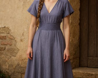 May include: A long, periwinkle-colored dress with a V-neck and short, scalloped sleeves. The dress features a cinched waist and a flowing skirt. The model has a long braid. The background includes a stone wall and a wooden door.