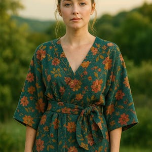 Puede incluir: Un vestido envolvente estilo kimono de color verde azulado con cuello en V y cinturón anudado. El vestido tiene diseños florales en naranja y óxido. Las mangas son hasta el codo. El fondo es un paisaje verde borroso.
