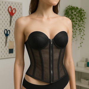 May include: A black strapless corset with a front zip. The corset has a structured bust and sheer mesh panels with vertical boning. The garment is designed to provide a fitted silhouette.
