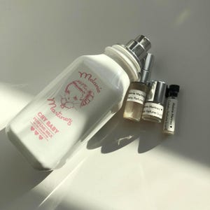 May include: A white perfume bottle with a silver sprayer and the words "Melanie Martinez" and "Cry Baby Perfume Milk" in pink. Several small perfume vials are next to the bottle. The bottle has a baby illustration.