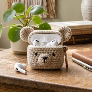 May include: A beige crocheted AirPod case shaped like a bear, with black embroidered eyes and nose. The case is open, revealing white AirPods inside. One AirPod rests beside the case on a wooden surface. A small metal clasp is attached to the case.