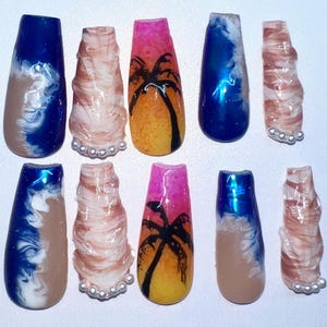 May include: A collection of ten artificial nails with diverse designs. Some nails display a blue and white wave pattern over a tan base. Others feature a sunset scene with a palm tree silhouette. Some nails have a pearl accent.