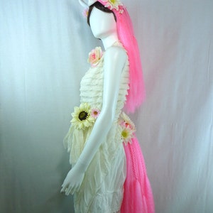 May include: A white dress with a ruffled skirt and a pink unicorn tail. The dress is decorated with white and yellow flowers. The model is wearing a white unicorn horn headband with pink and white flowers.