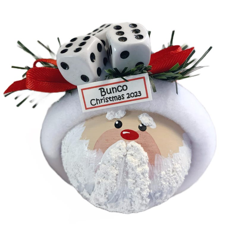May include: A round Christmas ornament featuring a painted Santa face with a white beard and hat brim. The top is decorated with green pine needles, a red ribbon, and three white dice. A small rectangular sign reads Bunco Christmas 2023.
