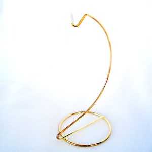 May include: A gold-colored metal display stand featuring a circular base and a curved, arching arm that extends upward. The top of the arm terminates in a small, white-tipped hook designed for suspending items, constructed from thin wire.