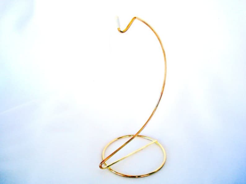 May include: A gold-colored metal display stand with a circular base and a curved, upward-reaching arm. The stand is designed to hold an ornament or other small decorative item. The stand is approximately 8 inches tall.
