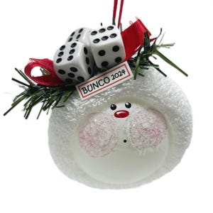 May include: A white snowman ornament with a red ribbon, green sprigs, and three dice. The ornament has pink cheeks, a red nose and mouth, and the text "BUNCO 2024" on a small white rectangle.
