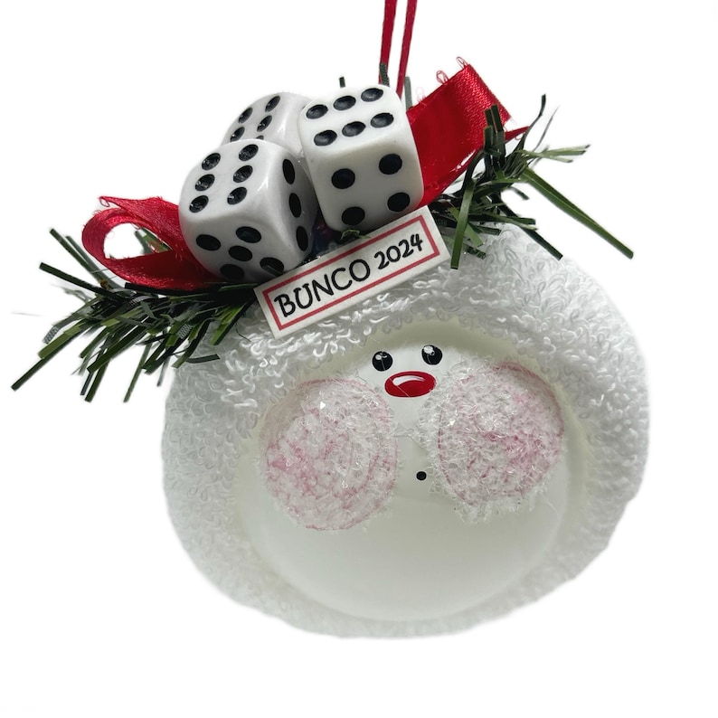May include: A white snowman ornament with a red ribbon, green sprigs, and three dice. The ornament has pink cheeks, a red nose and mouth, and the text "BUNCO 2024" on a small white rectangle.