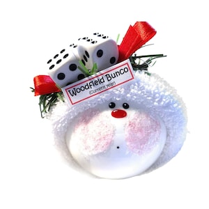 May include: A white ornament with a snowman design, featuring a red nose and pink cheeks. Two dice sit atop the snowman's head, along with a tag that reads "Woodfield Bunco (current year)". Red ribbon and green sprigs add festive detail.