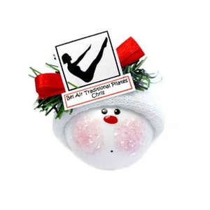 Puede incluir: Adorno de muñeco de nieve blanco con un gorro rojo y agujas de pino verdes. El adorno tiene una etiqueta blanca con el texto "Bel Air Traditional Pilates Chris".