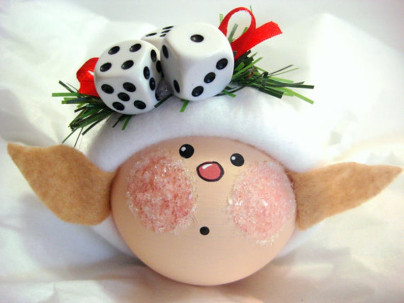 Puede incluir: Un adorno navide&ntilde;o caprichoso con una cara melocot&oacute;n, mejillas rosadas y orejas de fieltro. Est&aacute; rematado con tres dados blancos con puntos negros, cinta roja y ramitas verdes. El adorno est&aacute; sobre un fondo blanco.