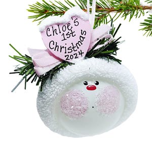 May include: A white Christmas ornament with a snowman face and a pink heart-shaped tag that reads "Chloe's 1st Christmas 2024". The ornament has a fluffy white hat and is decorated with a red nose and black eyes.