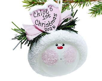 May include: A white Christmas ornament with a snowman face and a pink heart-shaped tag that reads "Chloe's 1st Christmas 2024". The ornament has a fluffy white hat and is decorated with a red nose and black eyes.