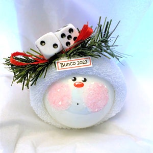 May include: A white snowman ornament with pink cheeks, a red nose, and black eyes. The ornament is topped with two dice, green sprigs, and a red ribbon. A small tag reads "Bunco 2022".